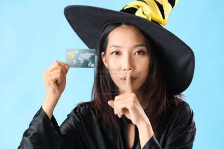 Young Asian woman dressed for Halloween as witch with credit card showing silence gesture on blue background, closeup