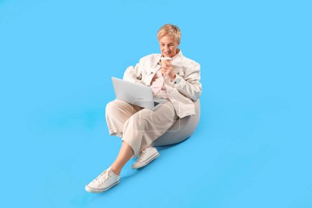 Téléchargez les photos : Femme mature programmeur avec ordinateur portable et tasse de café assis sur fond bleu - en image libre de droit