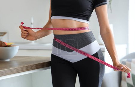Beautiful young woman measuring her waist in kitchen. Weight loss concept
