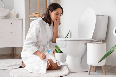 Téléchargez les photos : Jeune femme enceinte avec test souffrant de toxicose près de la cuvette des toilettes dans les toilettes - en image libre de droit
