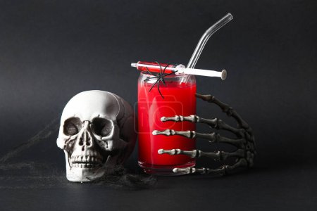 Glass of delicious red cocktail, skeleton hand, skull and syringe for Halloween celebration on black background