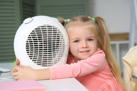 Photo for Cute little girl warming near electric fan heater at table in bedroom, closeup - Royalty Free Image