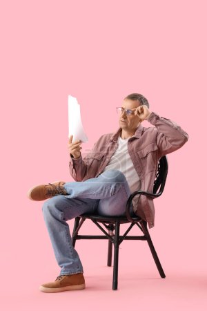 Téléchargez les photos : Mature acteur lecture film script dans chaise sur rose fond - en image libre de droit