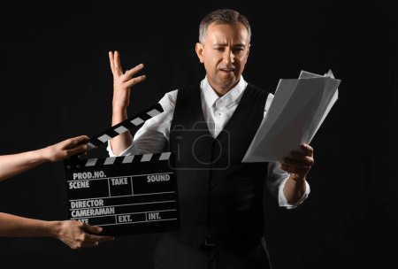 Téléchargez les photos : Mature acteur lecture script et mains avec clapperboard sur sombre fond - en image libre de droit