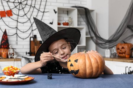 Téléchargez les photos : Petite fille mignonne habillée pour Halloween comme sorcière peinture citrouilles à table dans la cuisine - en image libre de droit