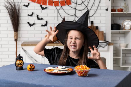 Téléchargez les photos : Petite fille mignonne habillée pour Halloween en sorcière avec des biscuits et des bonbons à table dans la cuisine - en image libre de droit