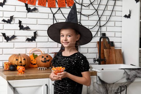 Téléchargez les photos : Petite fille mignonne habillée en sorcière avec des citrouilles et des bonbons dans la cuisine décorée pour Halloween - en image libre de droit