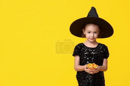 Téléchargez les photos : Petite fille mignonne habillée pour Halloween en sorcière avec bol de bonbons savoureux sur fond jaune - en image libre de droit