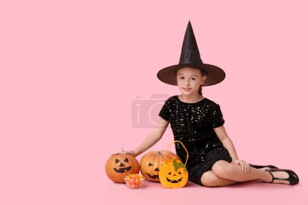 Téléchargez les photos : Petite fille mignonne habillée pour Halloween en sorcière avec sac, bonbons et citrouilles assis sur fond rose - en image libre de droit