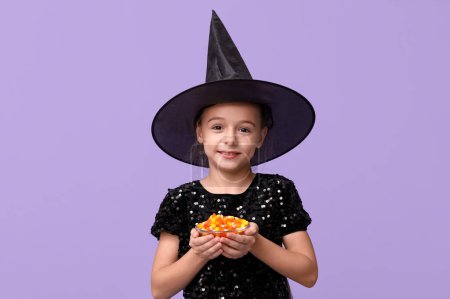 Téléchargez les photos : Petite fille mignonne habillée pour Halloween en sorcière avec bol de bonbons savoureux sur fond violet - en image libre de droit