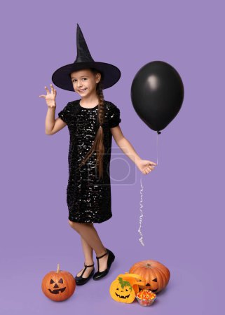 Téléchargez les photos : Petite fille mignonne habillée pour Halloween en sorcière avec des citrouilles, sac, ballon et bonbons sur fond violet - en image libre de droit