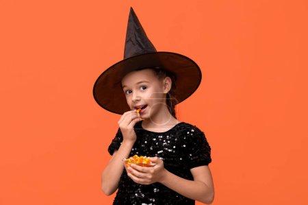 Téléchargez les photos : Petite fille mignonne habillée pour Halloween en sorcière avec bol de bonbons savoureux sur fond orange - en image libre de droit