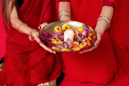 Photo for Beautiful mother with her daughter in sari holding candle and flowers on red background. Divaly celebration - Royalty Free Image