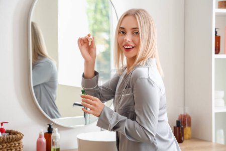 Téléchargez les photos : Jeune femme blonde appliquant du sérum capillaire près du miroir dans la salle de bain - en image libre de droit