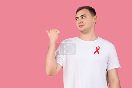 Foto de Joven guapo con cinta roja apuntando a algo sobre fondo rosa. Concepto del Día Mundial del SIDA - Imagen libre de derechos