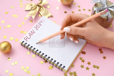 Woman writing PLAN 2024 in notebook and Christmas decor on pink background, closeup