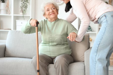 Photo for Young woman helping her mother with stick to get up from sofa at home - Royalty Free Image