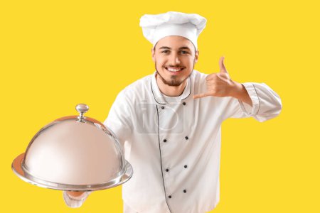 Téléchargez les photos : Chef masculin avec cloche montrant geste "appelez-moi" sur fond jaune - en image libre de droit