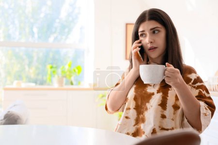 Téléchargez les photos : Jeune femme avec tasse de café bavardage par téléphone portable dans la cuisine - en image libre de droit