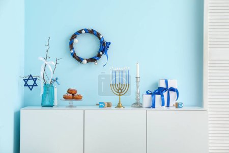 Téléchargez les photos : Armoire blanche avec menorah, sufganiyots, dreidels et coffrets cadeaux près du mur bleu. Hanoukka célébration - en image libre de droit