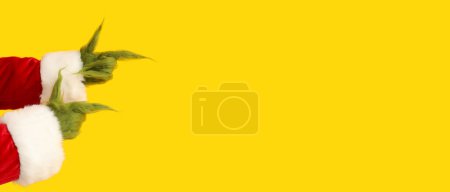 Photo for Green hairy hands of creature in Santa costume pointing at something on yellow background with space for text - Royalty Free Image