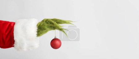 Green hairy hand of creature in Santa costume holding Christmas ball on light background with space for text