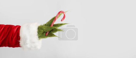 Photo for Green hairy hand of creature in Santa costume holding candy cane on light background with space for text - Royalty Free Image
