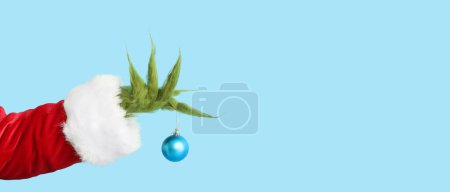 Main poilue verte de créature en costume de Père Noël tenant boule de Noël sur fond bleu clair avec espace pour le texte