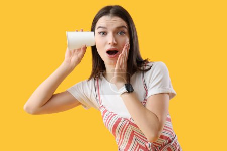 Photo for Shocked young gossip woman with cup trying to hear something on yellow background - Royalty Free Image