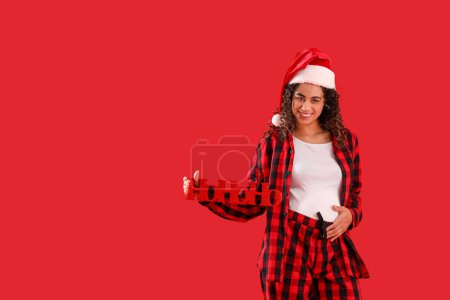 Téléchargez les photos : Femme afro-américaine enceinte dans le chapeau de Père Noël avec salutation de Noël sur fond rouge - en image libre de droit