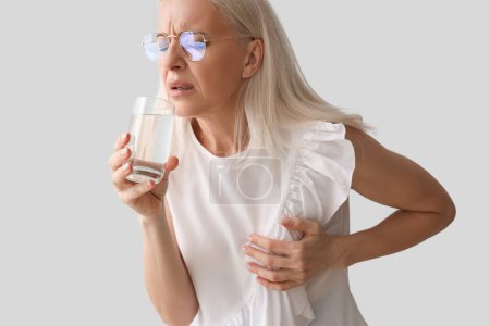 Téléchargez les photos : Femme mûre avec verre d'eau ayant une crise cardiaque sur fond gris, gros plan - en image libre de droit