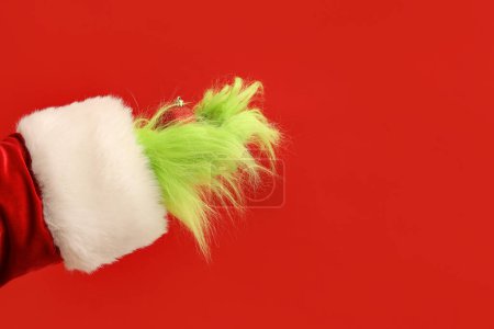 Téléchargez les photos : Main poilue verte de créature en costume de Père Noël avec boule de Noël sur fond rouge - en image libre de droit