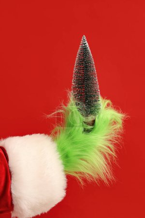 Téléchargez les photos : Main poilue verte de créature en costume de Père Noël avec sapin décoratif sur fond rouge - en image libre de droit