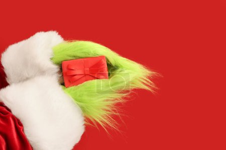 Foto de Verde manos peludas de criatura en traje de Santa con caja de regalo de Navidad sobre fondo rojo - Imagen libre de derechos