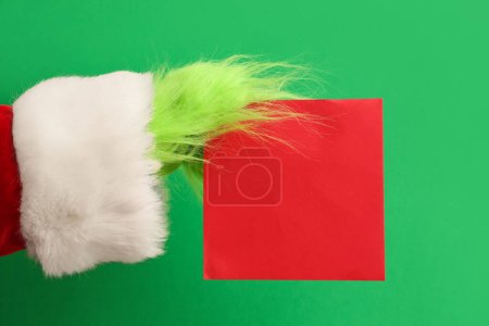 Photo for Green hairy hand of creature in Santa costume with blank card on green background - Royalty Free Image