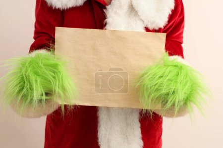 Photo for Green hairy hands of creature in Santa costume with blank paper sheet on white background - Royalty Free Image