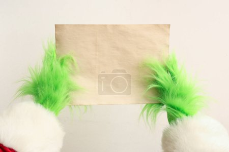 Téléchargez les photos : Mains poilues vertes de créature en costume de Père Noël avec feuille de papier vierge sur fond blanc - en image libre de droit