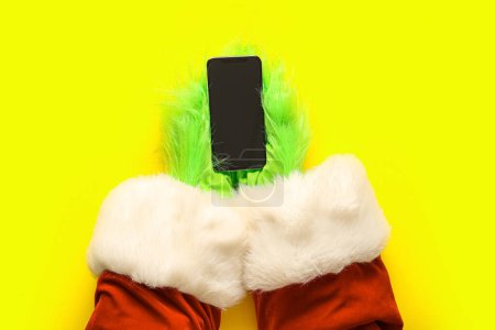 Téléchargez les photos : Mains poilues vertes de créature en costume de Père Noël avec téléphone portable sur fond jaune - en image libre de droit