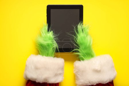 Photo for Green hairy hands of creature in Santa costume with tablet on yellow background - Royalty Free Image