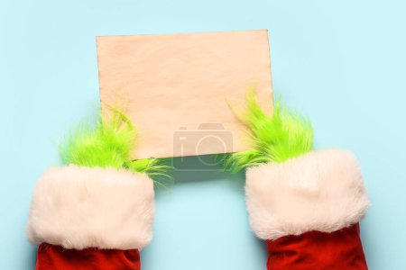 Photo for Green hairy hands of creature in Santa costume with blank paper sheet on blue background - Royalty Free Image