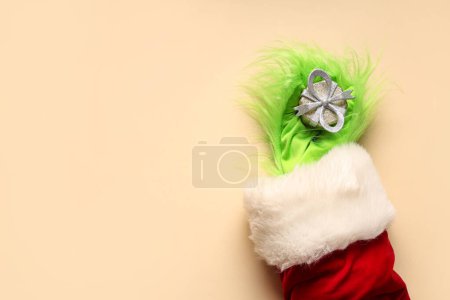 Photo for Green hairy hand of creature in Santa costume with Christmas gift box on beige background - Royalty Free Image