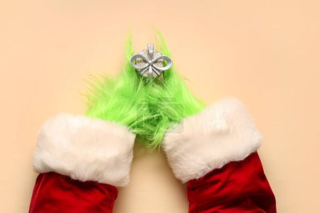 Téléchargez les photos : Mains poilues vertes de créature en costume de Père Noël avec boîte cadeau de Noël sur fond beige - en image libre de droit