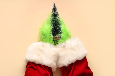 Photo for Green hairy hands of creature in Santa costume with decorative fir on beige background - Royalty Free Image