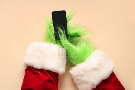 Photo for Green hairy hands of creature in Santa costume with mobile phone on beige background - Royalty Free Image