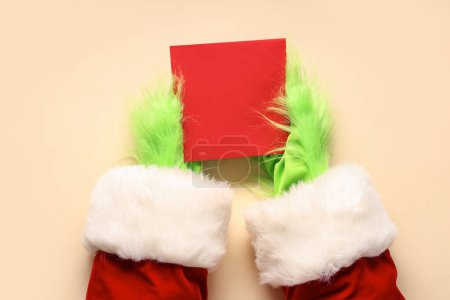 Téléchargez les photos : Mains poilues vertes de créature en costume de Père Noël avec carte blanche sur fond beige - en image libre de droit