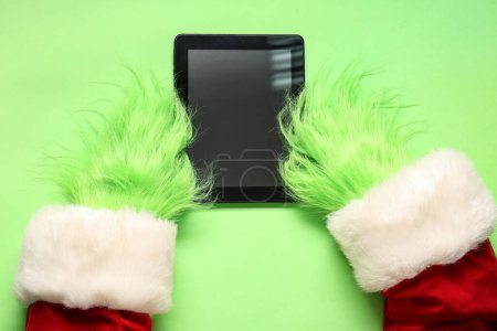 Photo for Green hairy hands of creature in Santa costume with tablet on green background - Royalty Free Image
