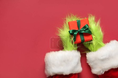 Photo for Green hairy hands of creature in Santa costume with Christmas gift box on red background - Royalty Free Image