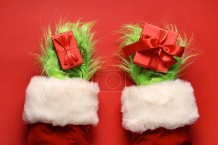 Photo for Green hairy hands of creature in Santa costume with Christmas gift boxes on red background - Royalty Free Image