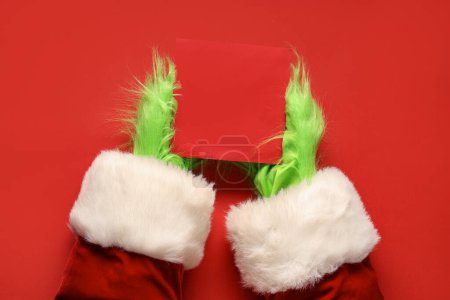 Téléchargez les photos : Mains poilues vertes de créature en costume de Père Noël avec carte blanche sur fond rouge - en image libre de droit