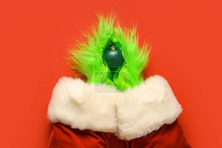 Téléchargez les photos : Mains poilues vertes de créature en costume de Père Noël avec boule de Noël sur fond rouge - en image libre de droit